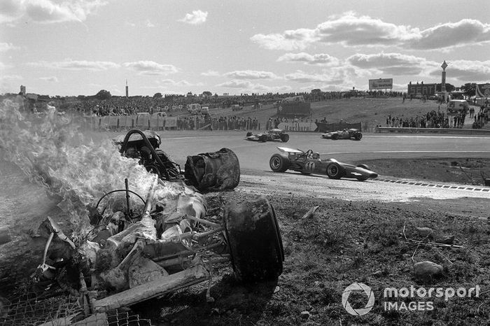 Bruce McLaren, McLaren M14A Ford, Graham Hill, Lotus 49C Ford, y Rolf Stommelen, Brabham BT33 Ford, pasan un un coche en llamas